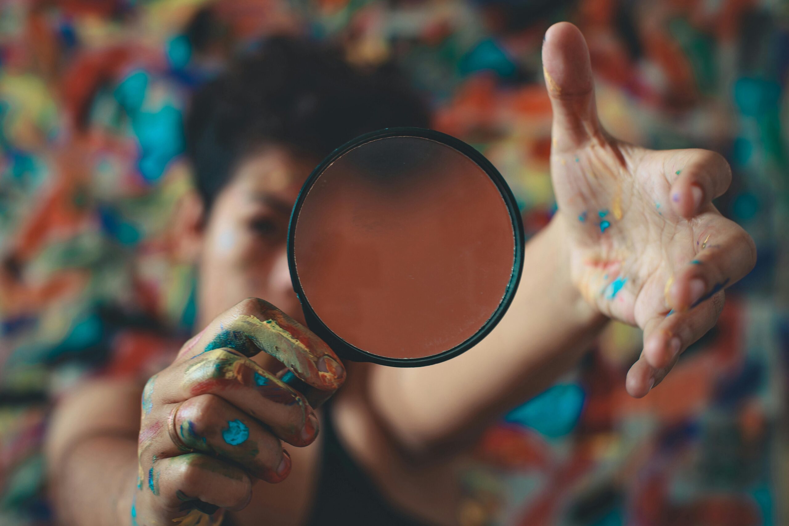 Artist with painted hands holding a lens in front of vibrant abstract background.